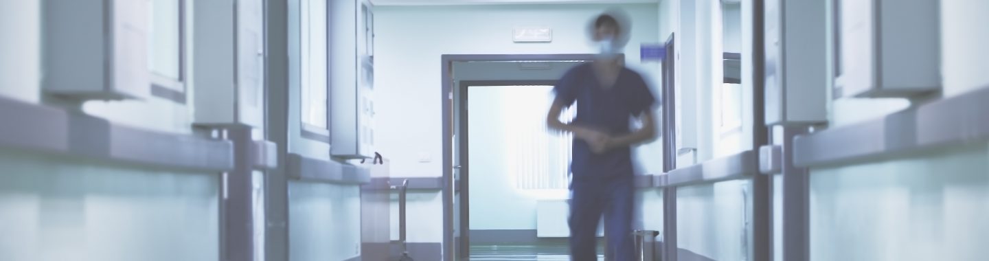 Person in scrubs with mask on walking forward down hospital hallway.